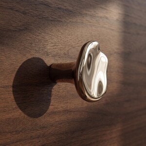 Peut inclure: Un bouton de meuble doré poli avec une forme organique irrégulière rappelant un liquide. Le bouton présente une surface métallique lisse et hautement réfléchissante, fixé sur une surface en bois brun foncé avec une ombre circulaire.