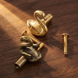 May include: Two brass cabinet knobs shaped like swans with detailed wings and necks. Each knob features a cylindrical base with a threaded hole for attachment. One knob is positioned upright, while the other lies on its side with a metal screw.