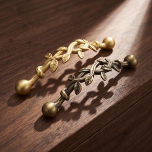 May include: Two metal cabinet pulls featuring a vine and leaf design with spherical ends. One pull has a bright gold finish, while the other has a darker, antique bronze finish. Both handles are shaped like intertwined branches with small leaves.