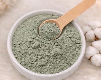 May include: A white bowl filled with green French clay, with a wooden scoop resting inside. The text "FRENCH GREEN CLAY" is displayed below, along with the words "DETOXIFYING • ABSORBENT • BALANCING".