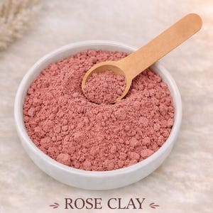 May include: A white bowl filled with pink rose clay powder, with a wooden scoop resting inside. The text "ROSE CLAY" is below the bowl, with the words "REVITALIZING • BALANCING • GLOW-BOOSTING" underneath.