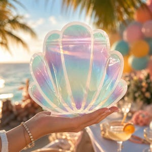 May include: Iridescent seashell-shaped bag with a zipper closure. The bag reflects a spectrum of colors, including blue, purple, and green. The bag is held in a hand, with a beach scene in the background. Perfect for beach-themed events.