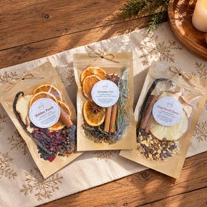 May include: Three clear pouches filled with dried fruit, spices, and herbs. The labels read Holiday Punch, Christmas Eve, and Gingerbread House. Each pouch is tied with twine and contains ingredients like orange slices, cinnamon sticks, and star anise.