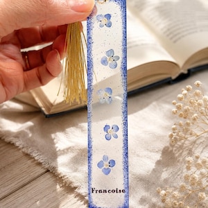 Peut inclure: Un marque-page rectangulaire avec une bordure bleue et une gland doré. Le marque-page présente des fleurs bleues incrustées et le nom "Francoise" en bas. Le fond est blanc avec de petits accents de paillettes.