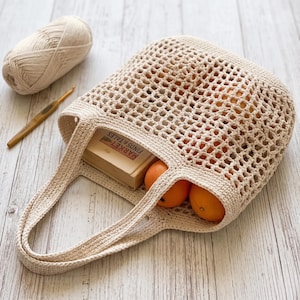 Peut inclure: Un sac fourre-tout en crochet beige avec un motif à mailles ouvertes, contenant deux fruits orange vif et un livre rectangulaire avec le texte MARKET FINDS & TALES sur la couverture. Une pelote de laine assortie et un crochet doré sont à côté.