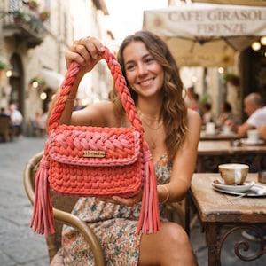 May include: A handmade crochet handbag in shades of coral, pink, and orange. The bag features a thick braided handle, a rectangular flap with a small wooden label reading "iforsale", and long decorative tassels hanging from both sides.