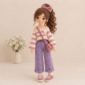 May include: A handcrafted crochet doll with brown curly hair styled in a high ponytail with a matching bow. The doll wears a striped off-the-shoulder sweater in pink and cream, purple wide-leg trousers, a small crossbody bag, and pink sneakers.