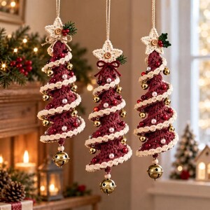 Op de afbeelding: Drie hangende kerstboomornamenten gemaakt van donkerrood gehaakt garen. Elke kegelvormige boom is omwikkeld met een crèmekleurige gehaakte slinger, versierd met kleine gouden belletjes, witte parels en rode kralen, bekroond met een crèmekleurige ster.