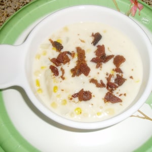 May include: A white bowl filled with creamy corn chowder topped with crispy bacon bits. The bowl is on a green and white patterned plate.