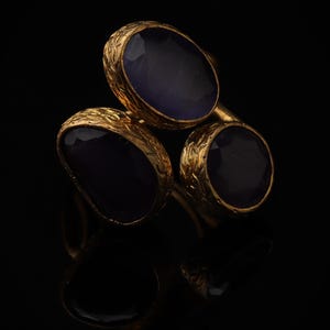May include: A gold-toned ring featuring three oval-shaped, deep purple cabochon stones. Each stone is set within a textured, metallic bezel with an intricate, etched pattern along the outer edges, creating a layered and dimensional appearance.