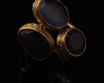 May include: A gold-toned ring featuring three oval-shaped, deep purple cabochon stones. Each stone is set within a textured, metallic bezel with an intricate, etched pattern along the outer edges, creating a layered and dimensional appearance.