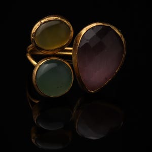 May include: A gold-toned ring featuring three distinct gemstones: a round olive-green stone, a round sage-green stone, and a larger, faceted, teardrop-shaped plum-colored stone. The metal band has a textured, organic finish and a warm yellow hue.