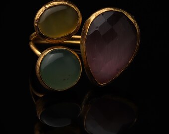 May include: A gold-toned ring featuring three distinct gemstones: a round olive-green stone, a round sage-green stone, and a larger, faceted, teardrop-shaped plum-colored stone. The metal band has a textured, organic finish and a warm yellow hue.