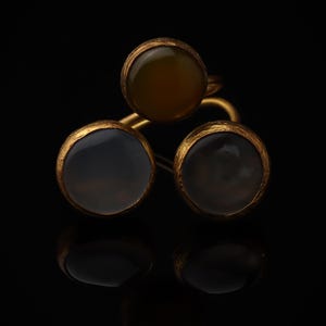 Könnte beinhalten: Ein goldener Ring mit drei kreisförmigen Fassungen, die glatte, durchscheinende Steine in Bernstein-, Grau- und Dunkelbrauntönen enthalten. Das Metallband und die Fassungen haben eine texturierte, matte Oberfläche und sind dreieckig angeordnet.
