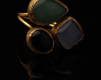 May include: A gold-toned ring featuring three distinct, irregularly shaped cabochon stones set in textured metal bezels. The stones include a dark green teardrop shape, a smooth black oval, and a translucent grey square-like stone.