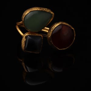 May include: A gold ring featuring three distinct stones set in textured metal settings. The stones include a teardrop-shaped green stone, a square black stone, and a circular reddish-brown stone, all arranged on a thin gold band.