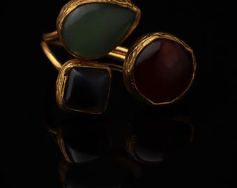 May include: A gold ring featuring three distinct stones set in textured metal settings. The stones include a teardrop-shaped green stone, a square black stone, and a circular reddish-brown stone, all arranged on a thin gold band.