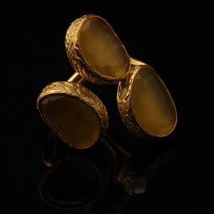 May include: A gold ring featuring three irregular, translucent amber-colored stones set in textured, organic-looking metal bezels. The band connects the three distinct stone settings, creating a layered appearance with a warm, metallic finish.