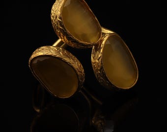 May include: A gold ring featuring three irregular, translucent amber-colored stones set in textured, organic-looking metal bezels. The band connects the three distinct stone settings, creating a layered appearance with a warm, metallic finish.