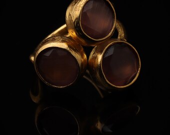 May include: A gold ring featuring three round, faceted, dark brown stones set in textured, matte gold bezels. The stones are arranged in a triangular cluster, with the metal band showing a hammered, organic finish throughout the design.