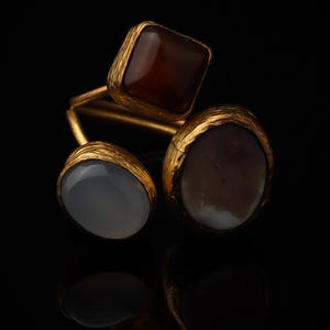 May include: A gold-toned ring featuring three distinct stones set in textured metal settings. The top stone is a square-shaped deep brown gem, while the two lower stones are oval-shaped, one being a translucent grey and the other a mottled brown.