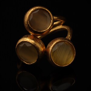 May include: A gold-toned ring featuring three round, faceted, translucent brownish-grey stones set in textured metal bezels. The stones are arranged in a cluster formation, with one stone positioned above two others on a continuous metal band.