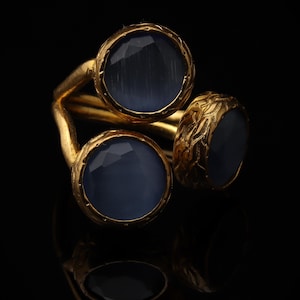 May include: A gold-toned ring featuring three round, faceted, slate-blue stones set in textured metal bezels. The band has a split design, with each stone held in a separate setting, creating a layered appearance against a dark background.
