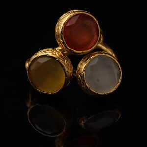 Könnte beinhalten: Ein goldener Ring mit drei runden, facettierten Edelsteinen in den Farben Dunkelorange, Senfgelb und durchscheinendem Weiß. Jeder Stein ist in eine texturierte, verzierte Goldfassung eingefasst, was ein Cluster-Design ergibt.
