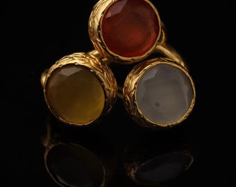 May include: A gold ring featuring three round, faceted gemstones in shades of deep orange, mustard yellow, and translucent white. Each stone is set within a textured, ornate gold bezel, creating a cluster design with a polished metallic finish.