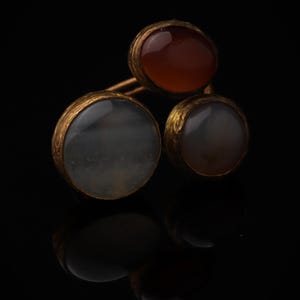 Könnte beinhalten: Ein Ring mit drei runden, in eine texturierte goldfarbene Metallfassung eingefassten Steinen. Die Steine umfassen einen durchscheinenden grauen, einen undurchsichtigen rotbraunen und einen halbtransparenten graubraunen Cabochon.