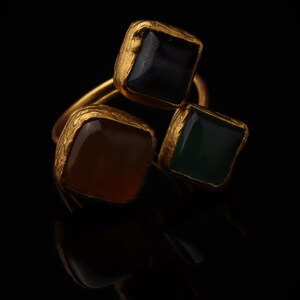 May include: A gold ring featuring three square-shaped stones in black, amber, and dark green. Each stone is set within a textured, irregular gold-toned metal frame, creating a cluster design with a polished finish on the stones and a rough edge.