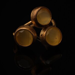 May include: A ring featuring three round, faceted, pale yellow stones set in a textured, dark bronze-toned metal band. The stones are arranged in a triangular cluster, with each stone held in a circular bezel setting that shows a rough, matte finish.