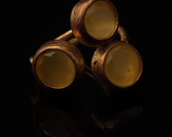 May include: A ring featuring three round, faceted, pale yellow stones set in a textured, dark bronze-toned metal band. The stones are arranged in a triangular cluster, with each stone held in a circular bezel setting that shows a rough, matte finish.