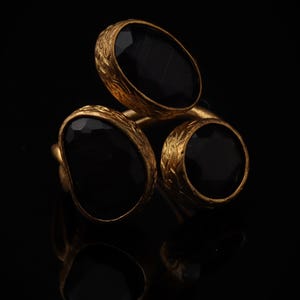 Könnte beinhalten: Ein goldener Ring mit drei facettierten schwarzen Steinen, die in texturierte, verzierte Goldfassungen eingefasst sind. Die Steine sind in einer Gruppe angeordnet, mit einem größeren ovalen Stein oben und zwei kleineren, runden Steinen darunter.