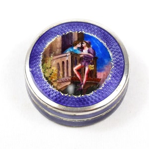 May include: A circular silver trinket box with a blue enamel border. The lid depicts a painted scene of two figures on a balcony, set against a blue sky. The box is suitable for jewellery or small keepsakes.