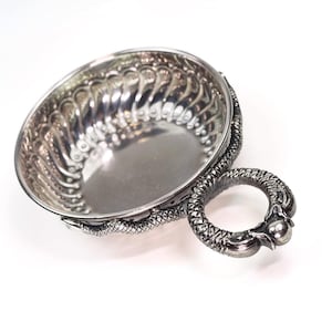 Può includere: Ciotola decorativa color argento con manico. La ciotola ha una forma ovale con un bordo traforato dettagliato. Il manico è a forma di anello, con un design strutturato. La ciotola è probabilmente in metallo.