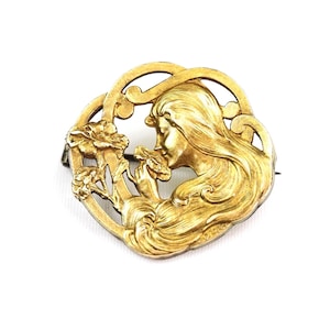 May include: A gold-toned Art Nouveau brooch featuring a woman with flowing hair, encircled by an ornate frame with floral accents. The woman is depicted in profile, delicately holding a flower. The brooch has a pin closure on the back.