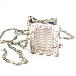 Puede incluir: Colgante de plata con forma de relicario cuadrado con tapa abierta, que revela un interior oscuro. El relicario está adornado con grabados florales y de volutas y cuelga de una cadena de plata. El relicario mide aproximadamente 2,5 cm cuadrados.