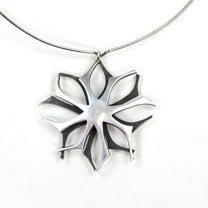 Op de afbeelding: Een zilverkleurige ketting met een hanger in de vorm van een gestileerde bloem. De hanger heeft een symmetrisch, open patroon met puntige, bloembladachtige vormen. De ketting heeft een dunne, zilverkleurige ketting.