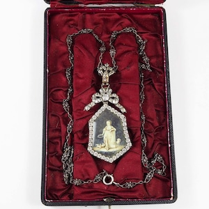 May include: An antique necklace with a pendant depicting a painted scene of a figure with a dog, framed by diamonds. The necklace is presented in a black box with a red velvet lining, highlighting the intricate details of the jewellery.