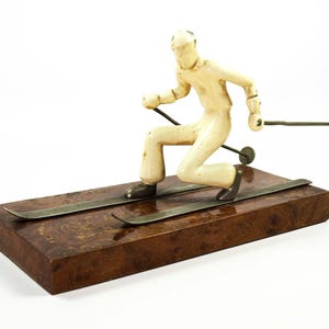Peut inclure: Figurine vintage d'un skieur en tenue crème, en pleine action. La figurine est montée sur une base rectangulaire en bois foncé avec des skis en métal. Le skieur tient des bâtons de ski en métal, capturant un moment de mouvement.