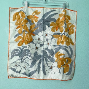 May include: Square silk scarf with a floral design. The scarf features a white background with a pattern of orange and white flowers, gray leaves, and an orange border. The scarf is hanging on a white hanger.