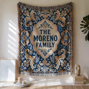 May include: A woven tapestry blanket featuring a central cream-colored cartouche with the text THE MORENO FAMILY in dark blue letters. The surrounding pattern consists of intricate blue, light blue, and tan floral scrollwork with fringed edges.