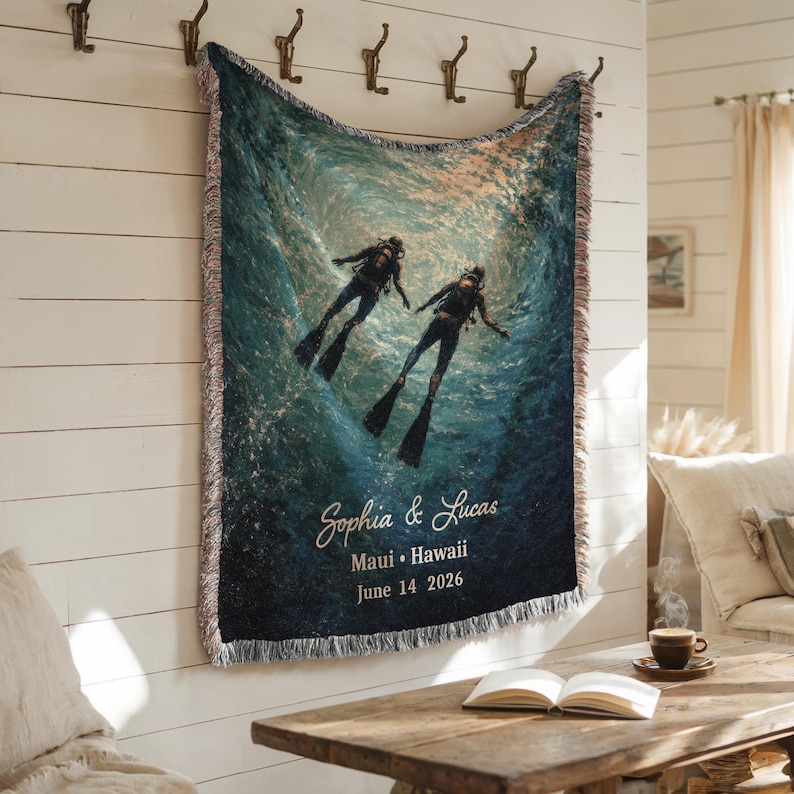 May include: Woven throw blanket featuring an underwater scene with two scuba divers. The blanket has a blue and teal color scheme with the names "Sophia & Lucas", location "Maui Hawaii", and date "June 14 2026" printed on it. The blanket is hanging on a white wall.