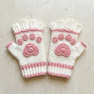 Può includere: Un paio di guanti senza dita all'uncinetto bianchi e rosa. Ogni guanto presenta un motivo a zampa rosa sul palmo e impronte più piccole sul dorso. I guanti hanno un bordo rosa e sono realizzati in filato morbido e strutturato.