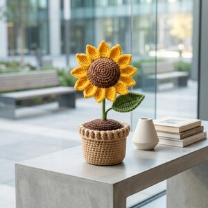 May include: A crocheted sunflower in a pot. The sunflower has yellow petals and a brown center. The pot is a light brown color and has a decorative edge. A green leaf is attached to the stem. The item is sitting on a gray surface.