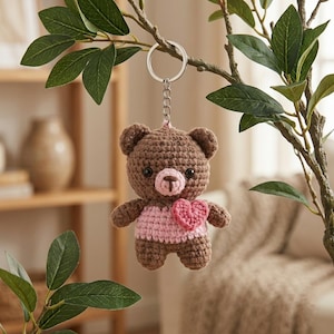 Peut inclure: Un porte-clés en forme d'ours en peluche marron crocheté, avec un cœur et un pull roses. L'ours est suspendu à un porte-clés argenté, avec un nez brun clair et des yeux noirs. L'ours est suspendu à une branche d'arbre avec des feuilles vertes.