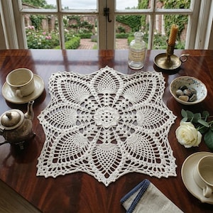 Puede incluir: Un tapete de ganchillo color crema con un diseño floral, colocado sobre una mesa de madera oscura. El tapete está rodeado por un juego de té, un pequeño cuenco de piedras, una vela y una rosa blanca. Una ventana con vistas al jardín está al fondo.