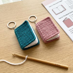 May include: Two crochet book keychains, one teal and one pink, with white pages. A wooden crochet hook and a crochet pattern are also visible. The keychains are on a light brown wooden surface.