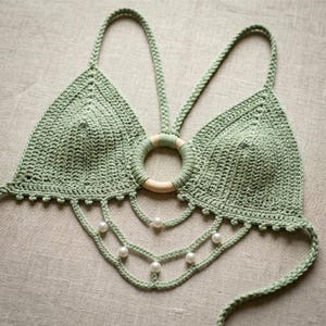 Può includere: Top all'uncinetto verde chiaro con design all'americana. Il top presenta un dettaglio con anello in legno ed è adornato con perle finte. Il lavoro all'uncinetto è visibile, mostrando la consistenza e l'artigianalità del capo.